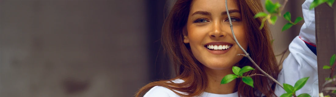 Dentist Feeding Hills MA Teeth Whitening Focused a woman with a spray tan is leaning against a shrub while looking off to the left and smiling